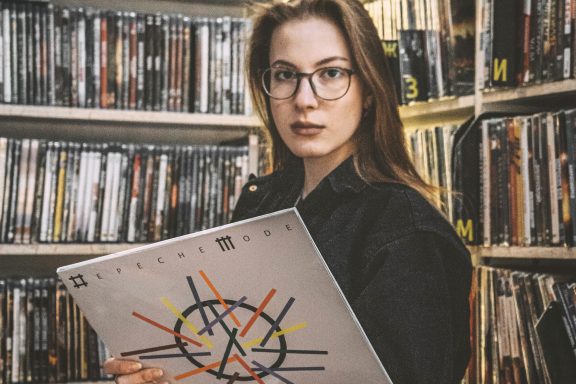 Frau mit Brille hält eine Schallplatte vor einem Regal voller CDs.